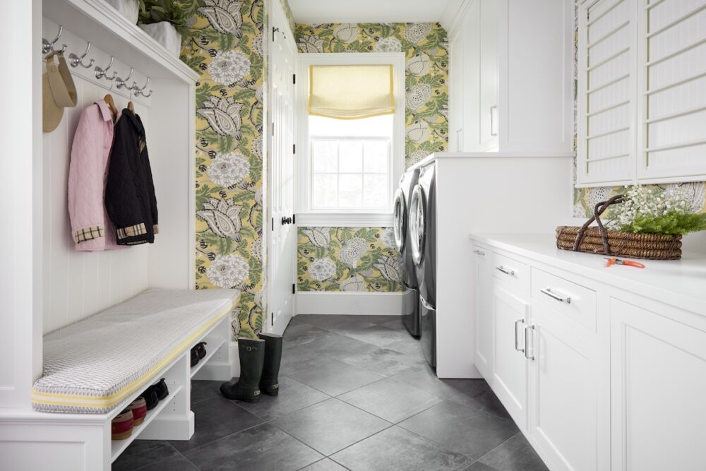 A Thibaut print is used as wallpaper in the mud/laundry room with plenty of welcome storage. A built-in bench holds shoes topped with a cushion in small geometric Schumacher print bordered in a yellow Samuel & Sons tape trim.