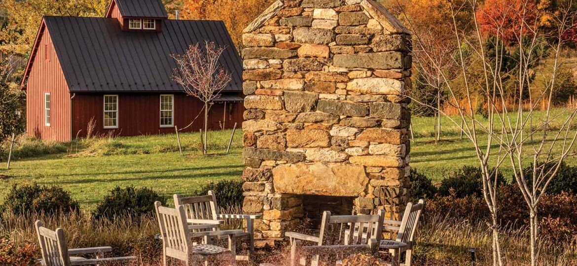 Stacked stone fireplace with seating area in garden in front of red barn. Autumn-dried hydrangeas and grasses border seating area.