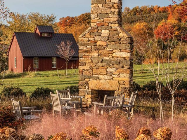 Stacked stone fireplace with seating area in garden in front of red barn. Autumn-dried hydrangeas and grasses border seating area.