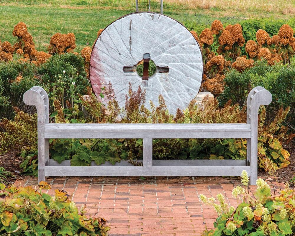 Wooden garden bench in front of an antique millstone framed by a bank of hydrangeas.