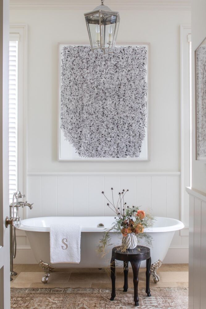 Clawfoot bathtub in primary bath with painting by Mark Fox on wall, autumn flower arrangement on stool beside tub.