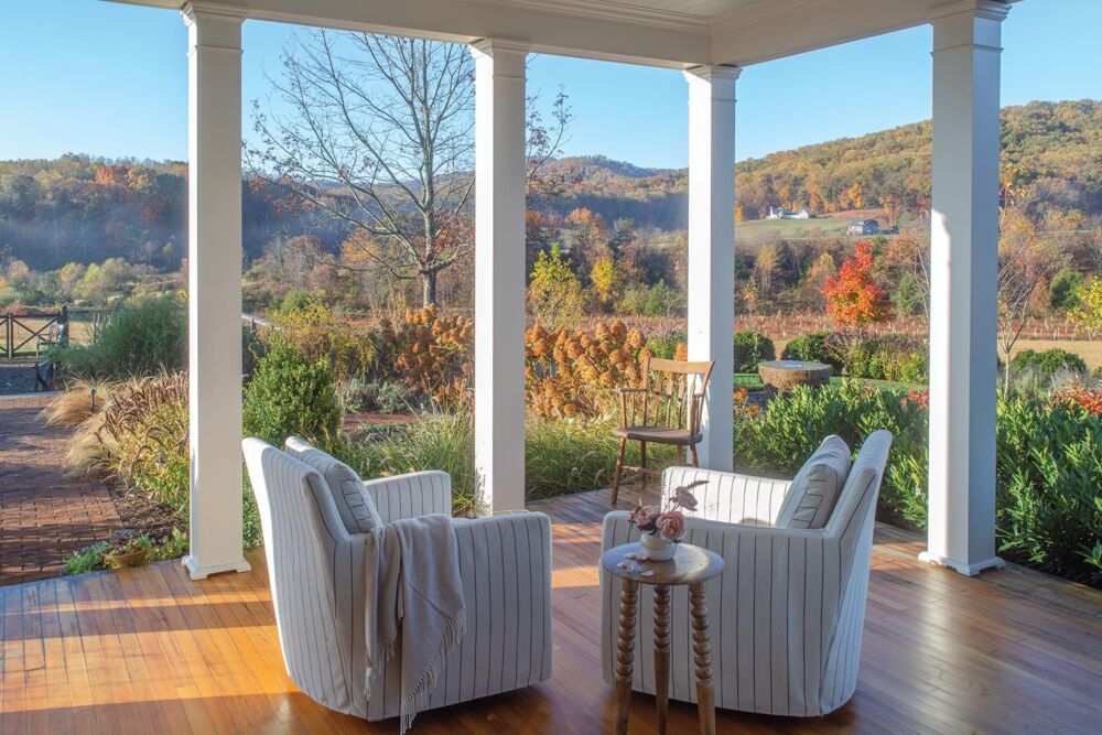 Pair of chairs on a columned side porch with views of the garden and hills beyond in fall color.
