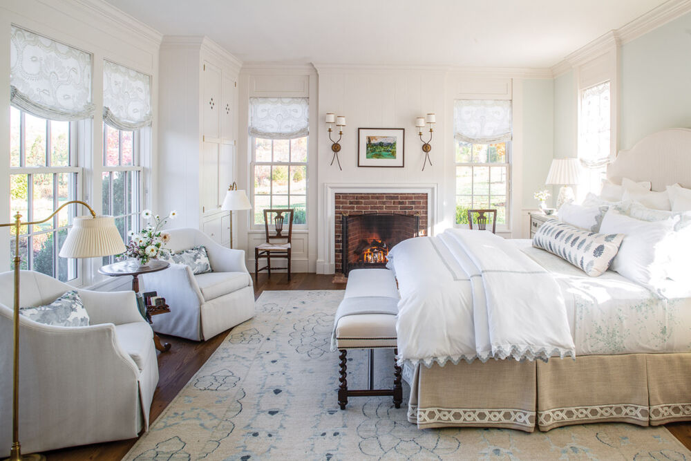 Primary bedroom with white linens and touches of blue, windows on three sides, fireplace with pair of sconces and painting over.
