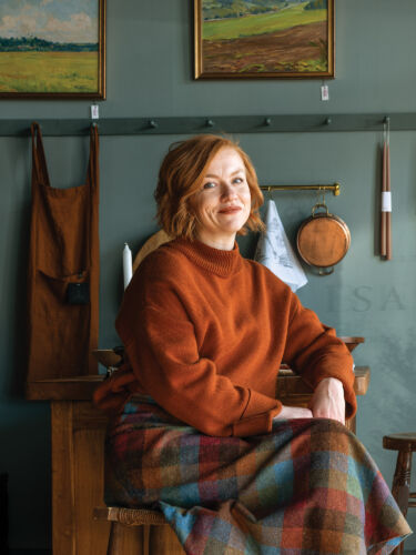A red-haired woman sits in her dark teal storefront in an orange sweater and flannel skirt.