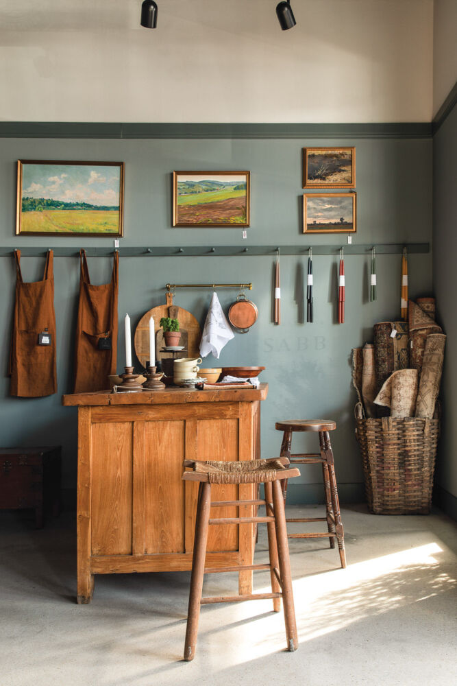 A teal-grey wall holds rust colored aprons, paintings, and candles.