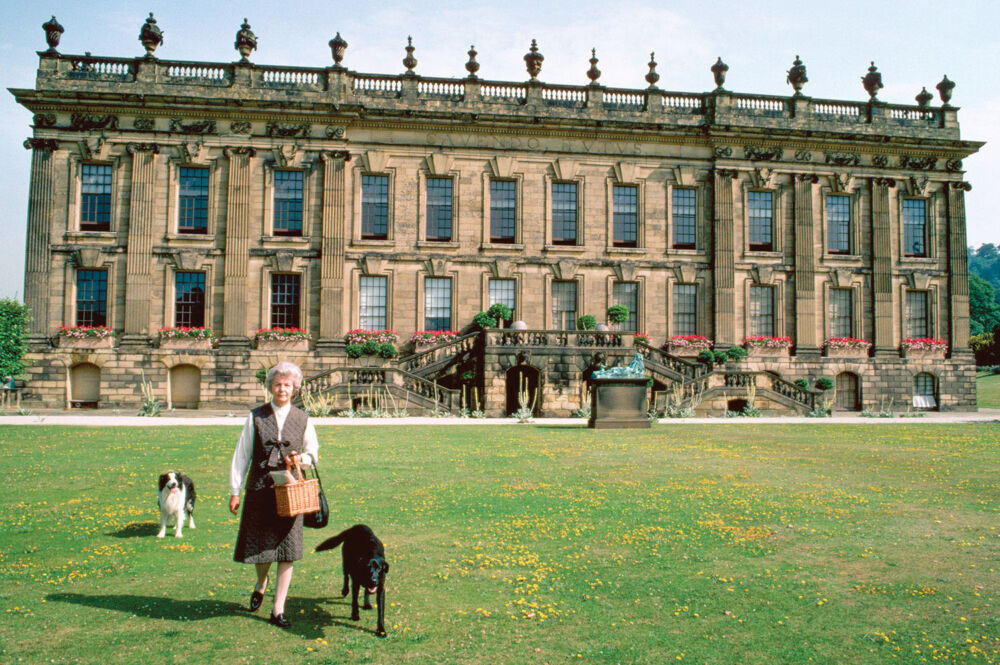 The Duchess of Devonshire strides along her grounds with her two dogs and her palace behind her.