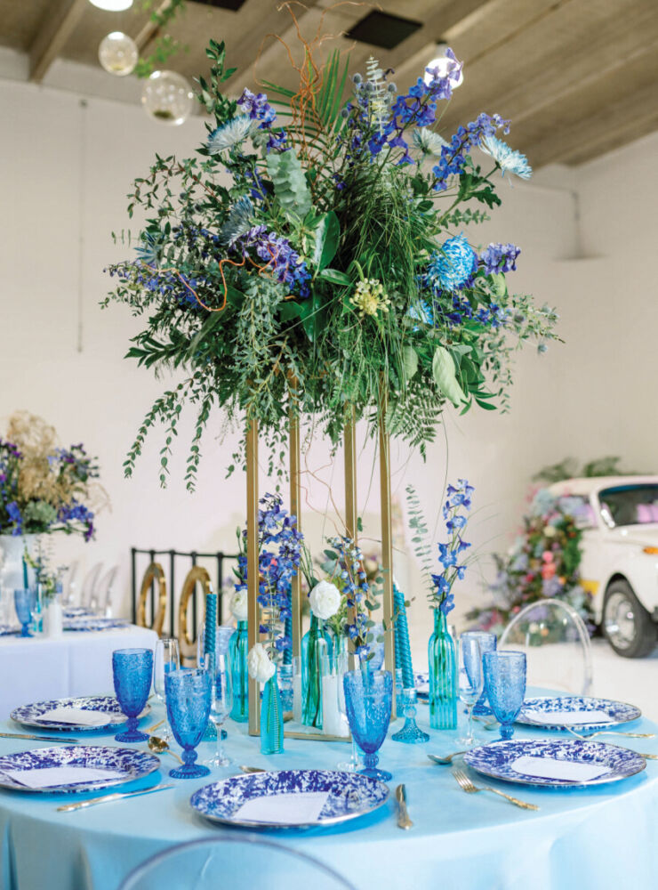 A bushel of greenery and wildflowers tower over a blue table.