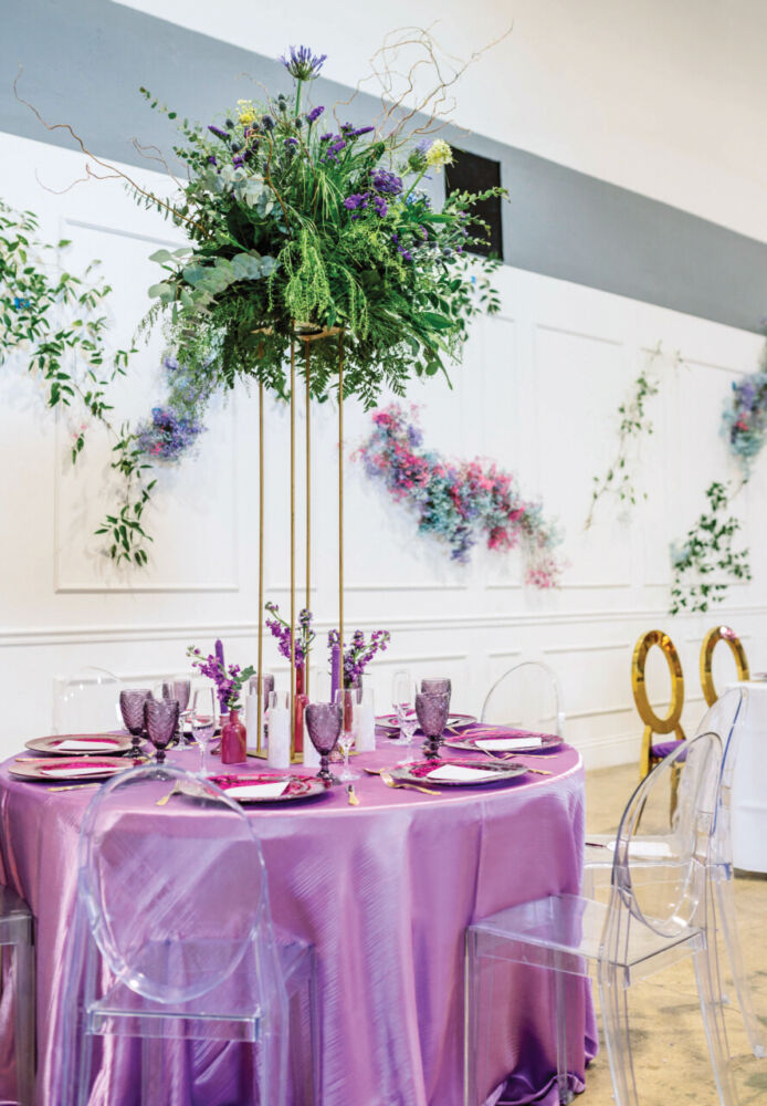Wild flowers burst out of a tall stilted vase on a purple tablecloth.