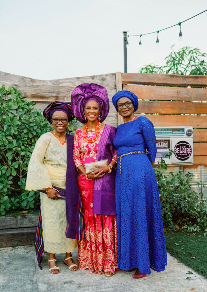 Three women stand smiling, dressed in traditional Nigerian outfits.
