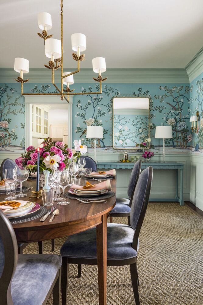 Dining room with blue floral de Gournay wallpaper, and centerpiece of pinkish purple roses on table. Streamlined lighting fixture above the table.