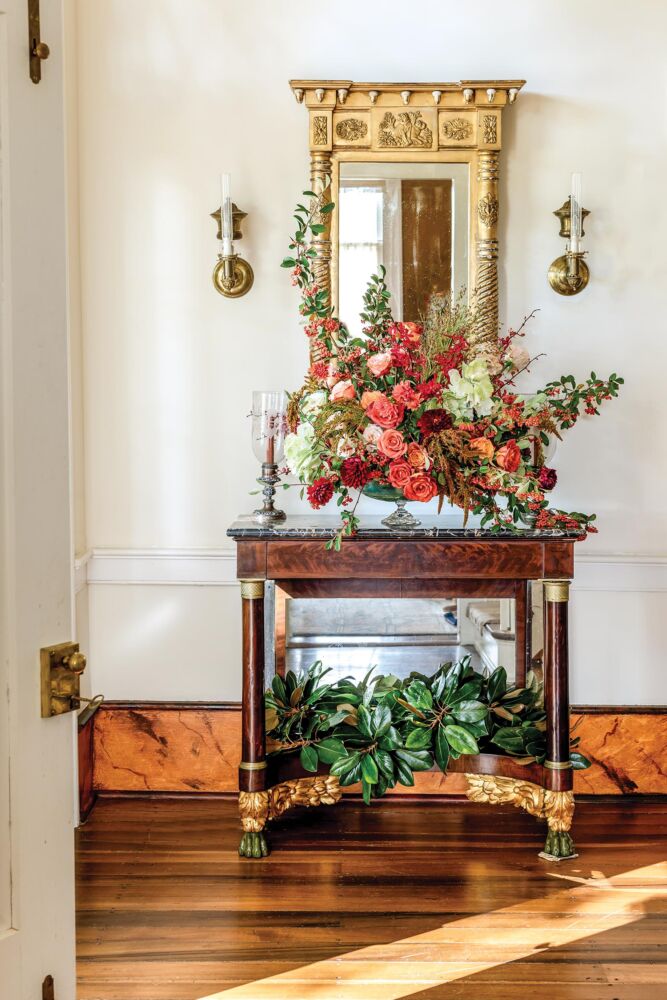 A bountiful fall bouquet with coral roses sits on top of an antique wooden table in front of a gold rimmed mirror in a sunlit entryway.