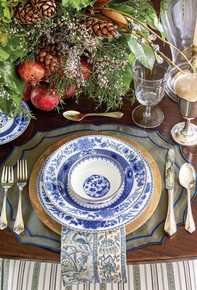 Blue and white china lays on top of a grey placemat with silverware and flowers surrounding.