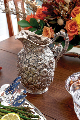 A silver floral engraved pitched stands on a wooden table next to an orange bouquet of flowers.