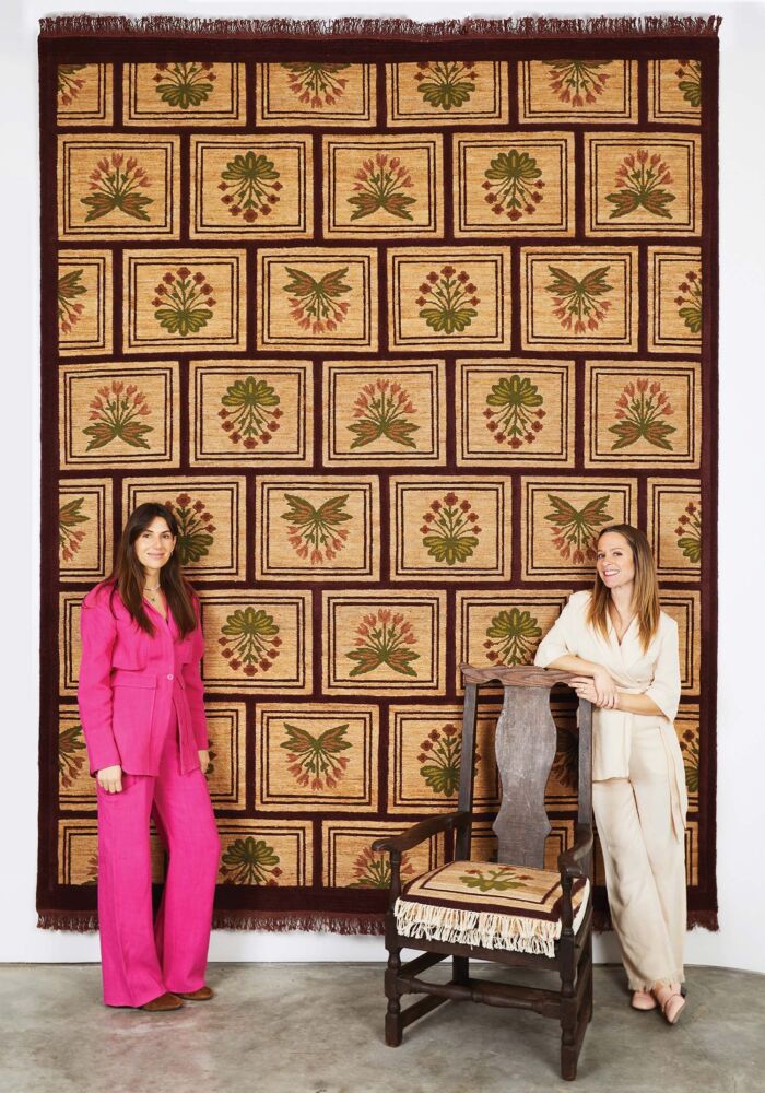 A floral rug hangs on the wall behind two women.