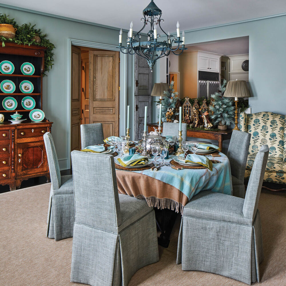 Grey linen covered dining chairs sit around a teal velvet table in a teal painted room.