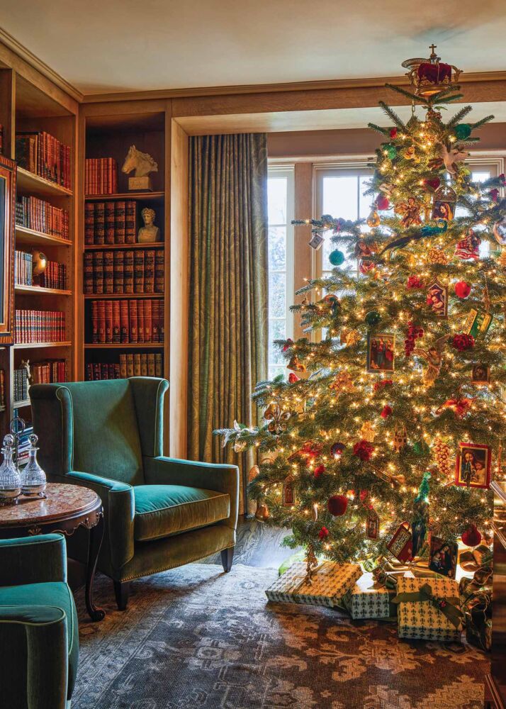 A lit and heavily ornamented Christmas tree stands in a sitting room next to two velvet emerald chairs.