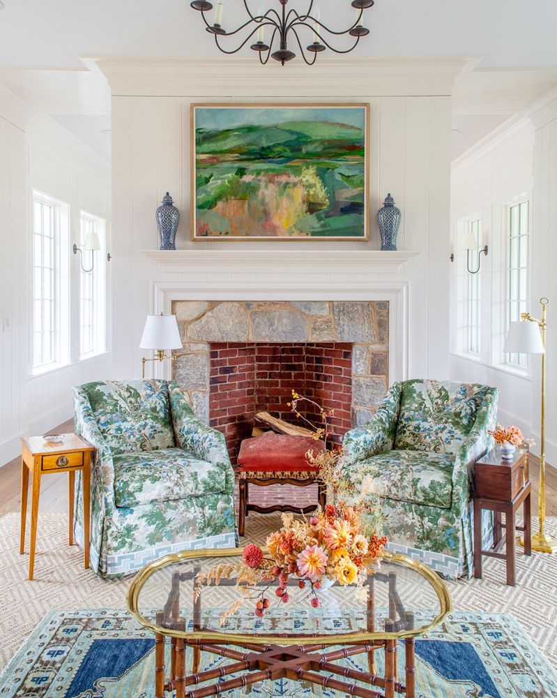 Floral upholstered chairs are centered around a painted landscape above a fireplace.