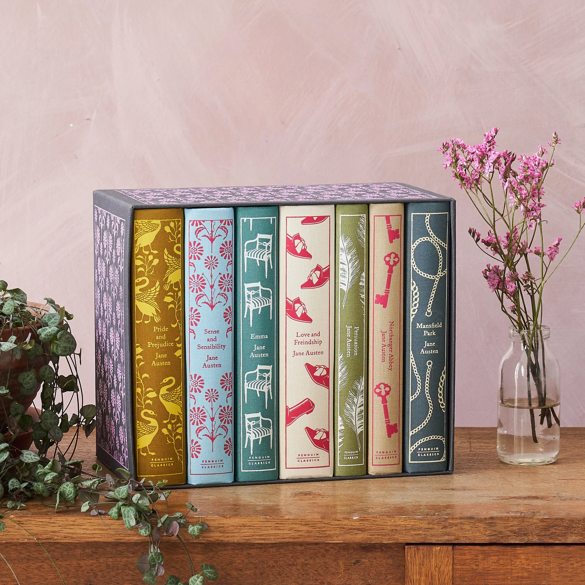 7 clothbound books sit on a wooden table next to flowers.