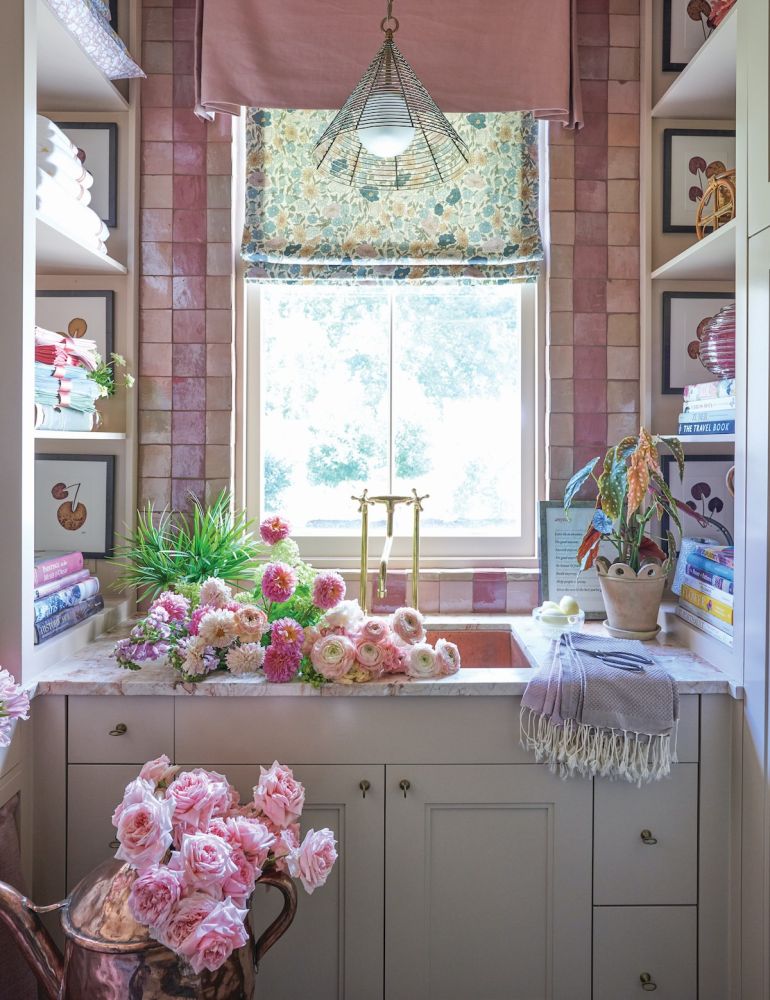 Laundry room with pink tile on wall over sink, Liberty fabric Roman shade on window, shelves flanking sink counter. Sink is filled with pink roses and other flowers.