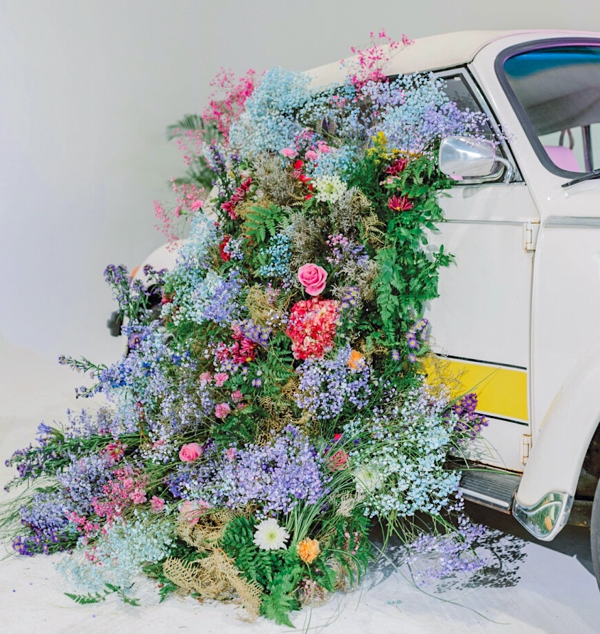 A waterfall of baby’s breath, delphinium, chrysanthemums, agapanthus, purple asters, carnations, roses, hydrangeas, goldenrod, dried fern, green fern, Italian ruscus, and grasses spill out of a 1969 Volkswagen bug.