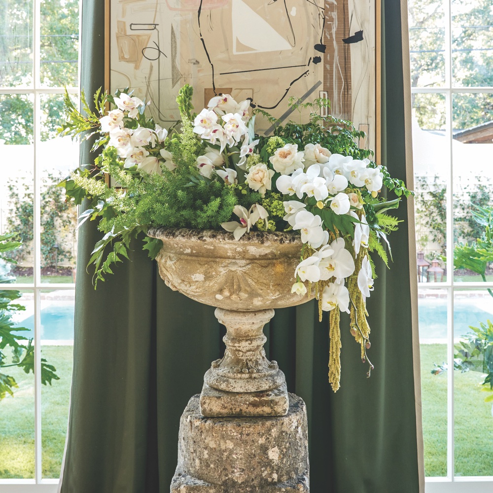 White orchids and roses spill out of an aged concrete container in front of large windows.