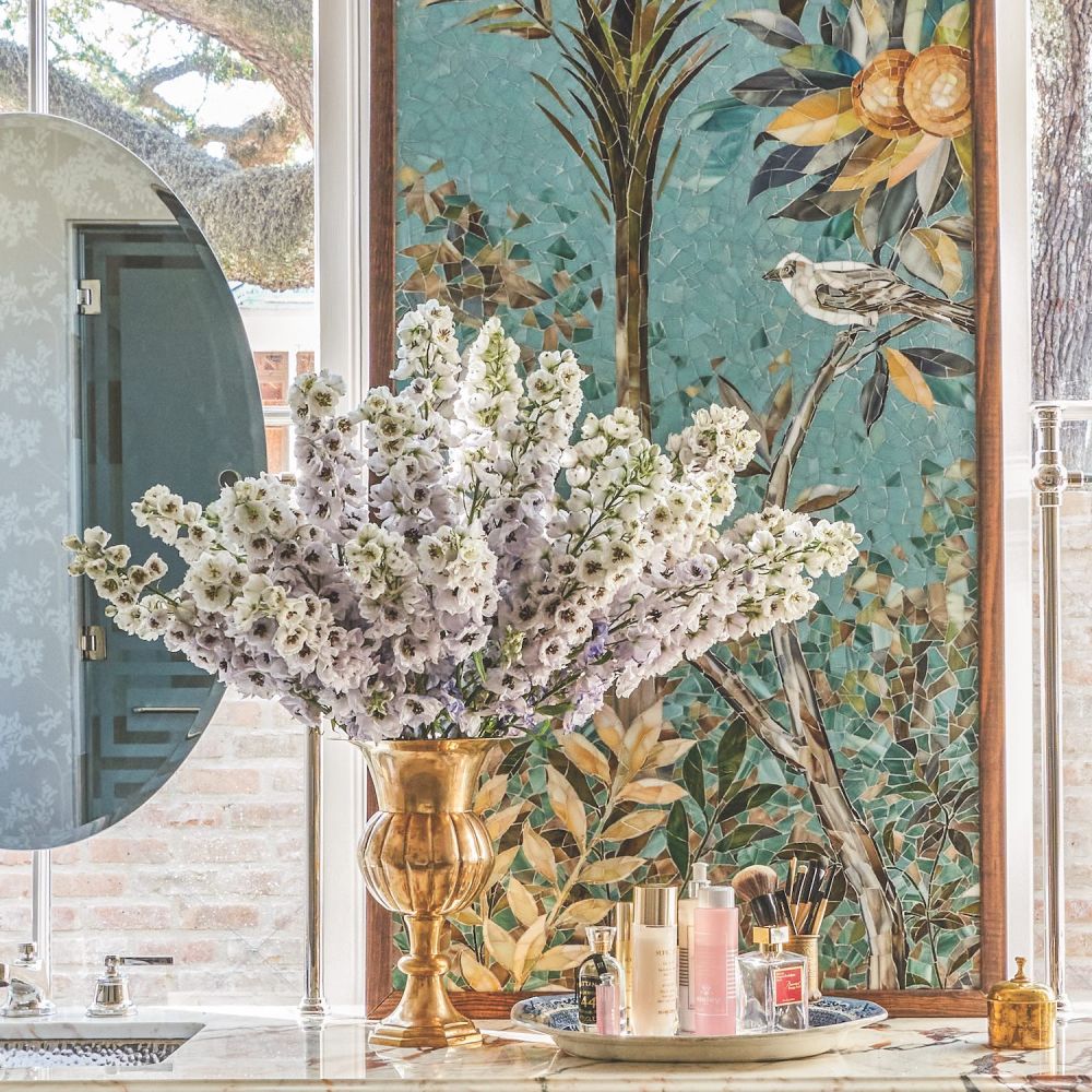 Brass vase of lilac-colored double-flowering delphinium on vanity in front of bird and flower mosaic.