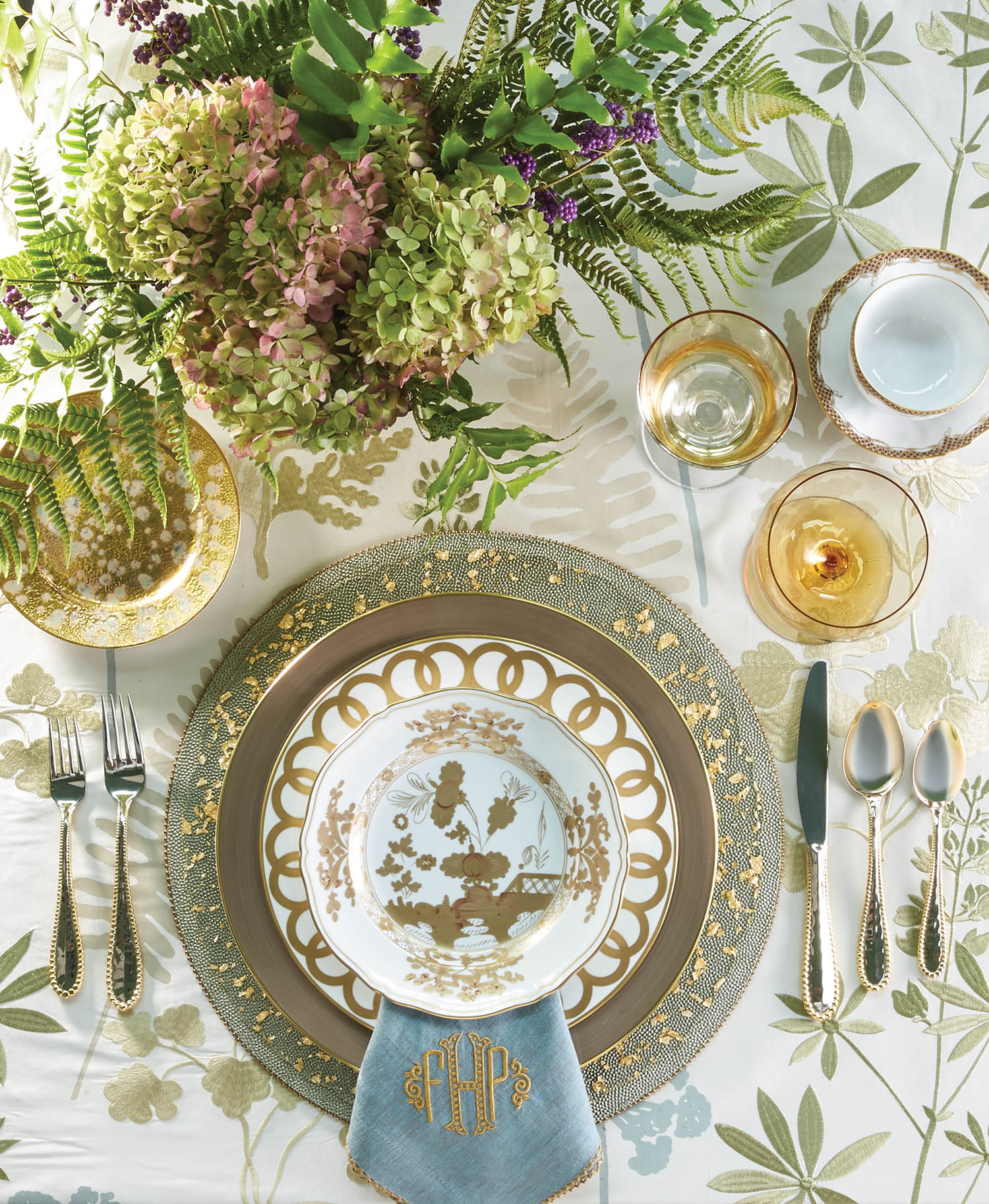 Table setting in tones of gold and green with arrangement of hydrangeas and fern fronds.