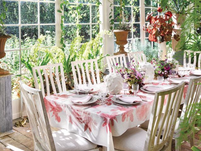 A long table with a pink and white floral tablecloth sits in a sunroom decorated with hanging greenery.