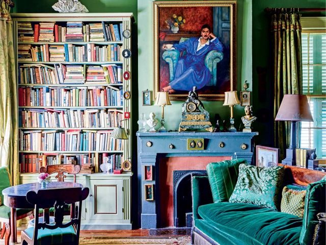 A green room with a portrait hanging over a fireplace, a stuffed bookshelf, and a velvet couch.