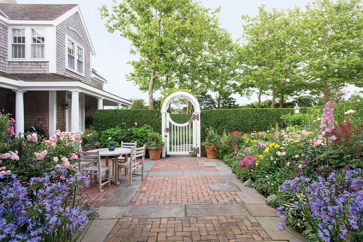 A blooming garden leads to a white gate outside a cedar-shake house.