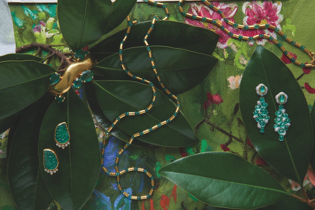 Emerald jewelry that includes a pair of large emerald stone earrings, a gold and emerald necklace lay across dark green leaves