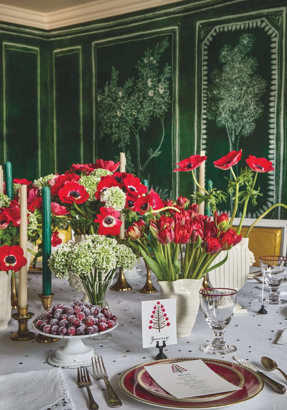 A holiday table is dressed with a white tablecloth, crystal glassware, red and gold plates, a plate of sugared cranberries, a vase of red tulips and a vase of red anemones, and brass candle holders with tapered candles in them.