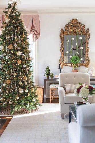 A decorated Christmas tree sits in a living area next to an antique mirror on the wall that is adorned with ornaments hanging from ribbon from the top of the mirror. Miniature Christmas trees sit on a short table in front of the mirror.