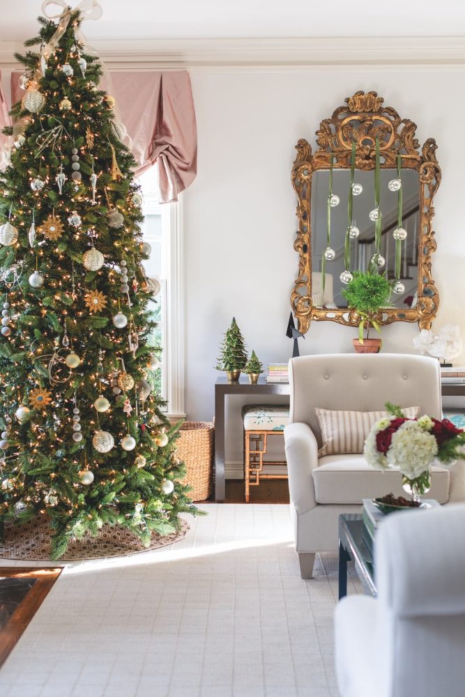 A decorated Christmas tree sits in a living area next to an antique mirror on the wall that is adorned with ornaments hanging from ribbon from the top of the mirror. Miniature Christmas trees sit on a short table in front of the mirror.
